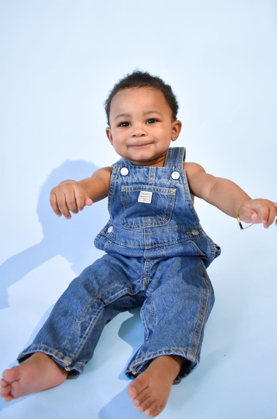 A baby in denim overalls sitting on a pale blue studio backdrop in Las Vegas