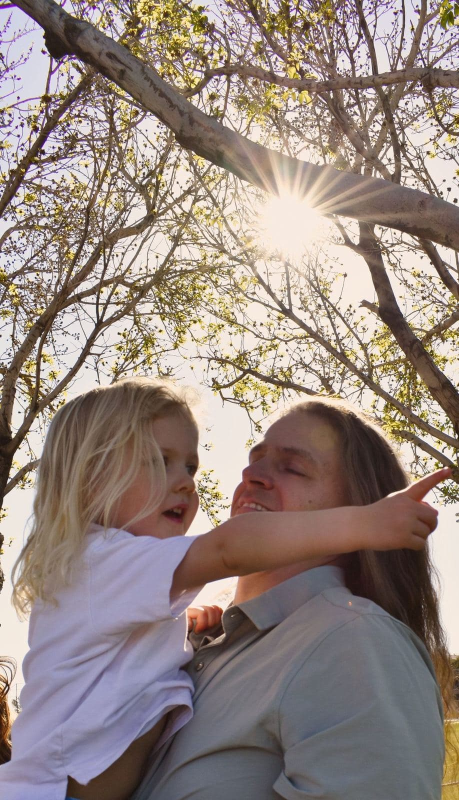 A father holding his young daughter as she reaches upward, sunlight breaking through the trees overhead in Las Vegas, NV