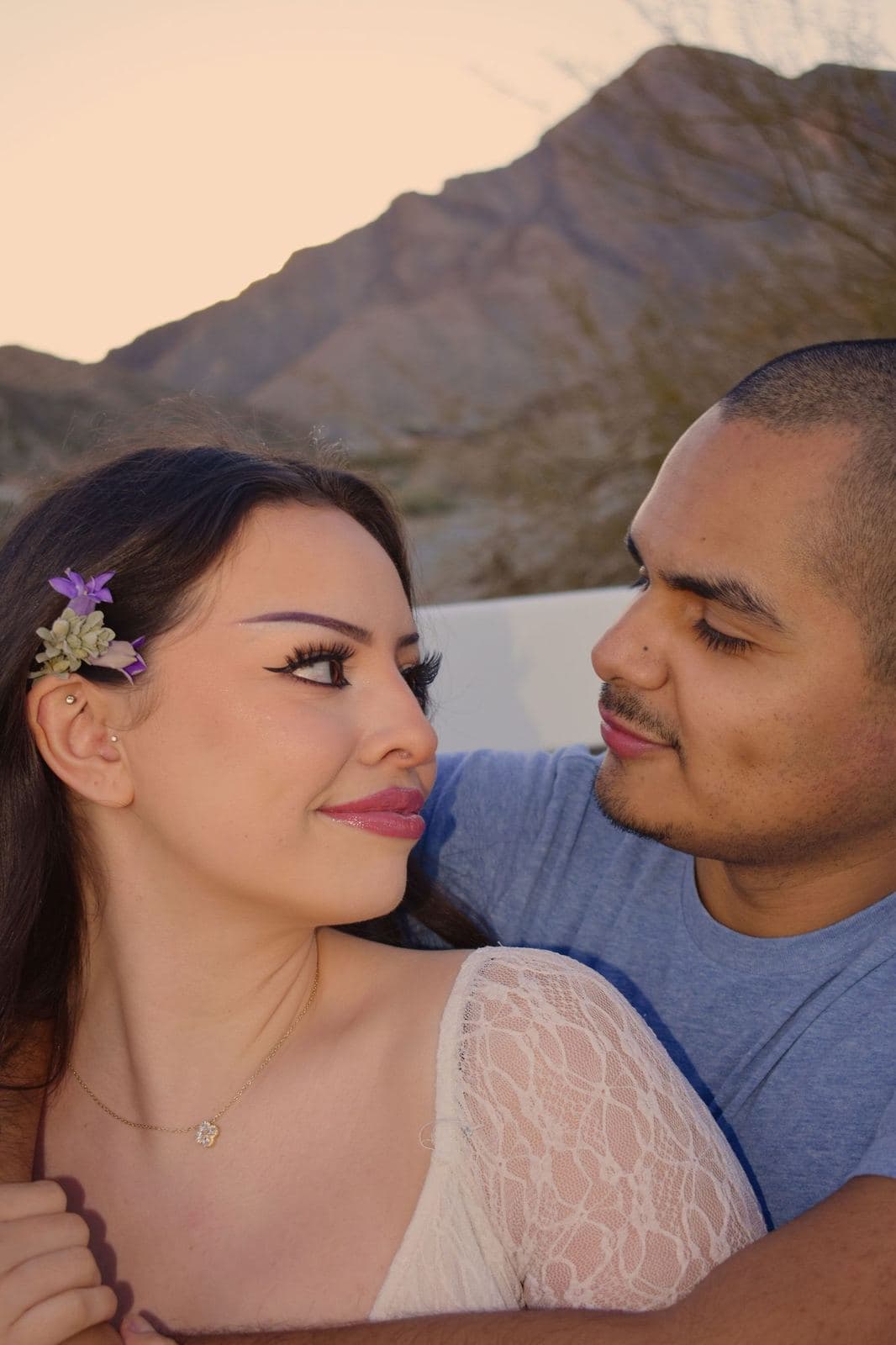 A close portrait of a couple looking at each other, a small purple flower in her hair and a desert mountain behind them near Las Vegas, NV