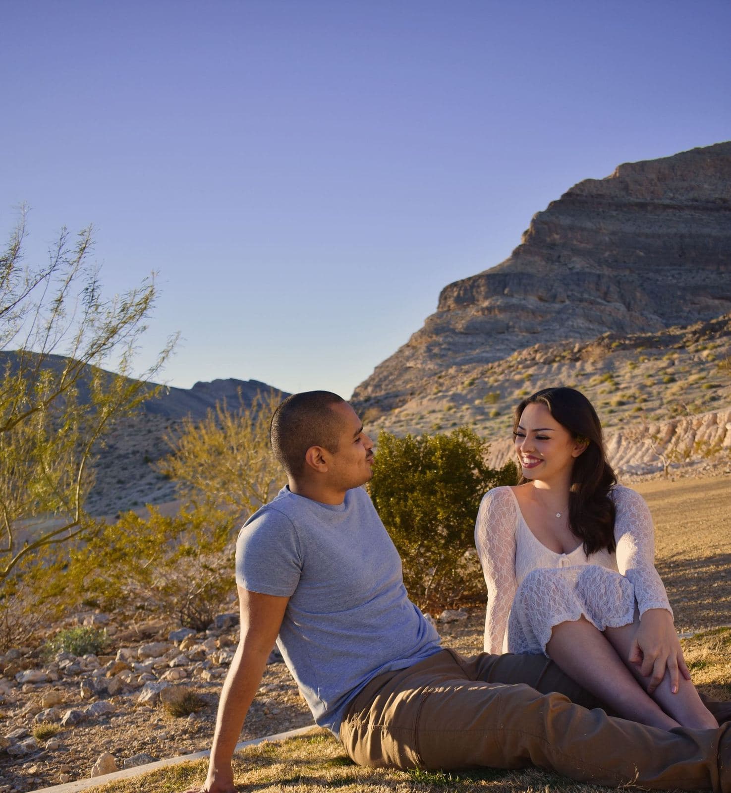 A couple sitting together on the ground below red desert cliffs at golden hour near Las Vegas, NV