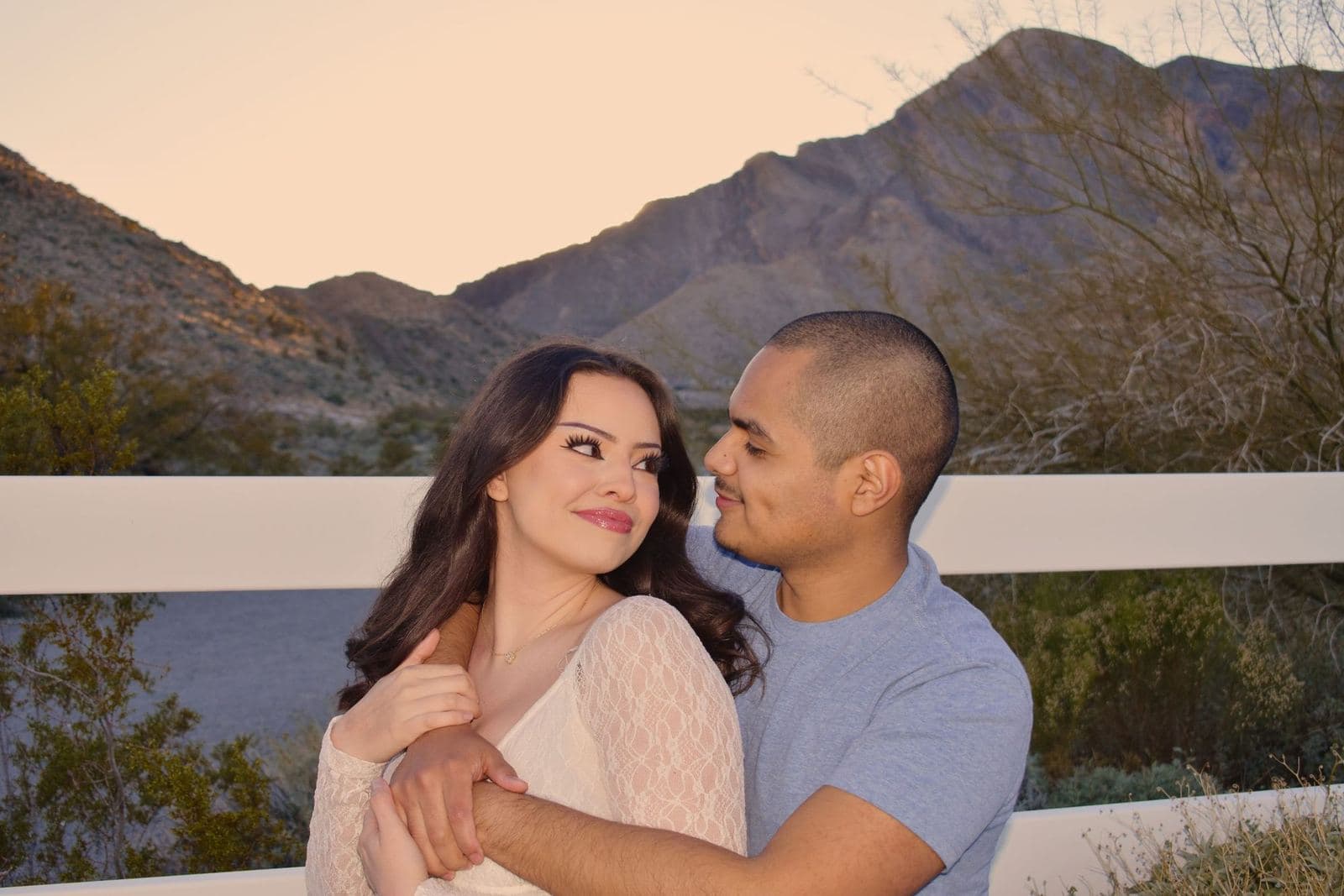 A couple looking at each other beside a white ranch fence below a desert mountain at sunset near Las Vegas, NV