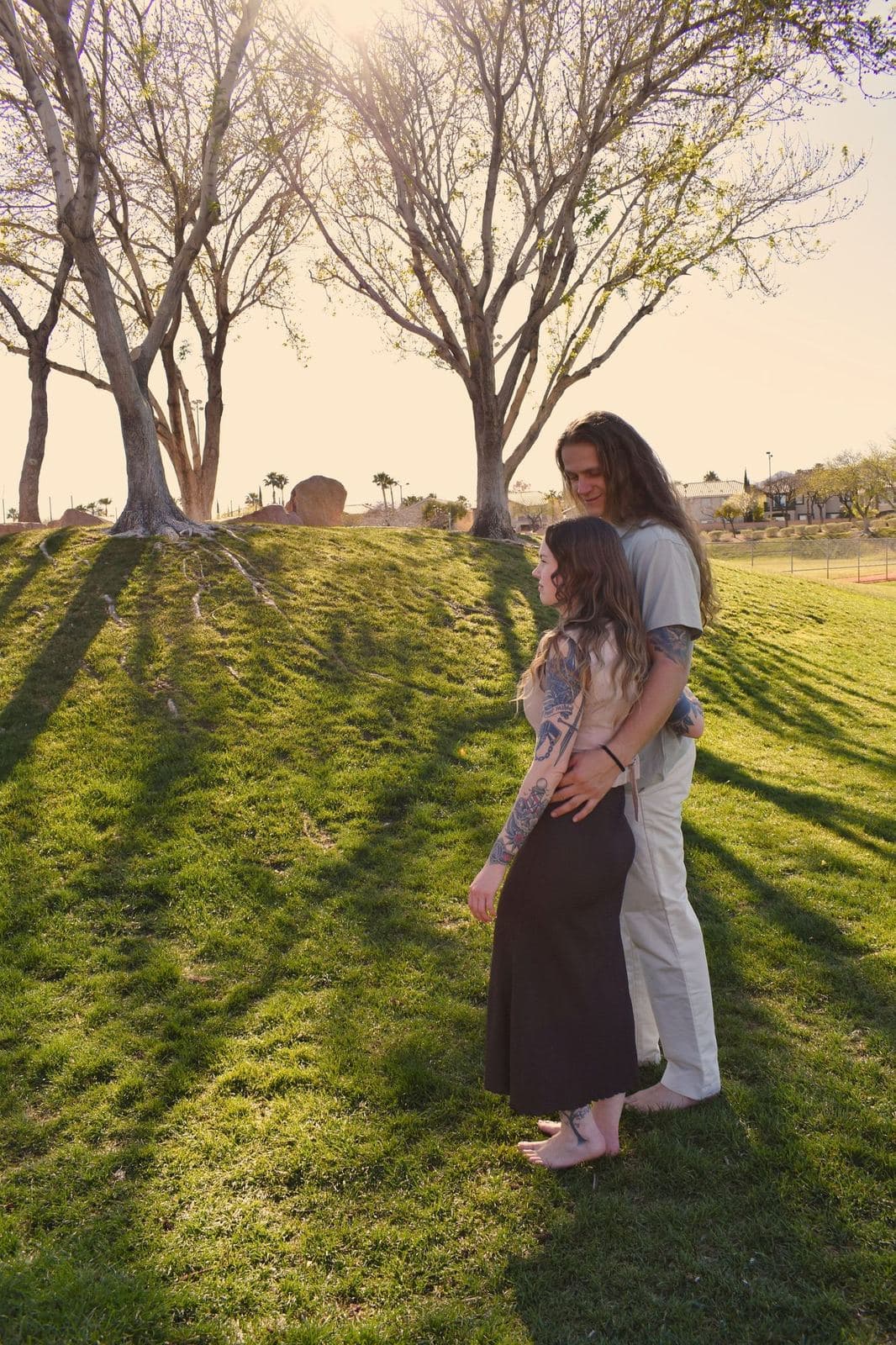 A couple standing together on a park lawn beneath bare trees, long shadows across the grass at sunset in Las Vegas, NV