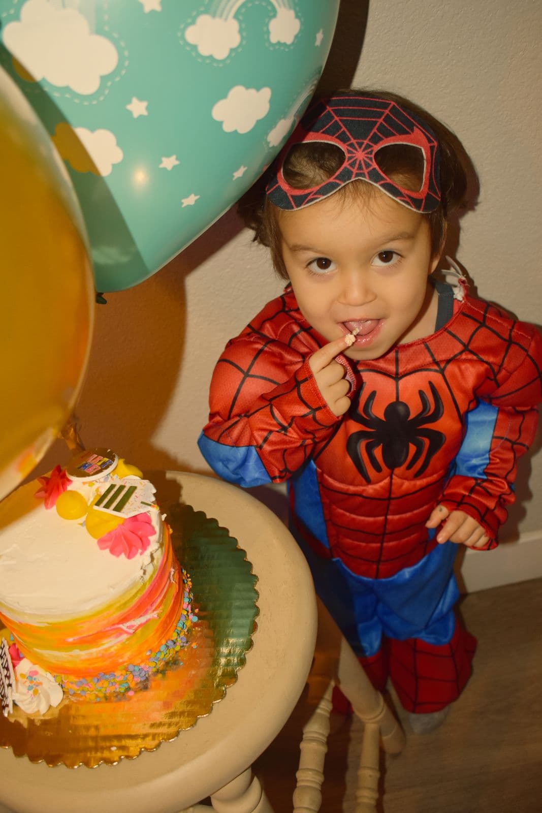 A young boy in a superhero costume grinning beside a bright layered birthday cake, Las Vegas, NV