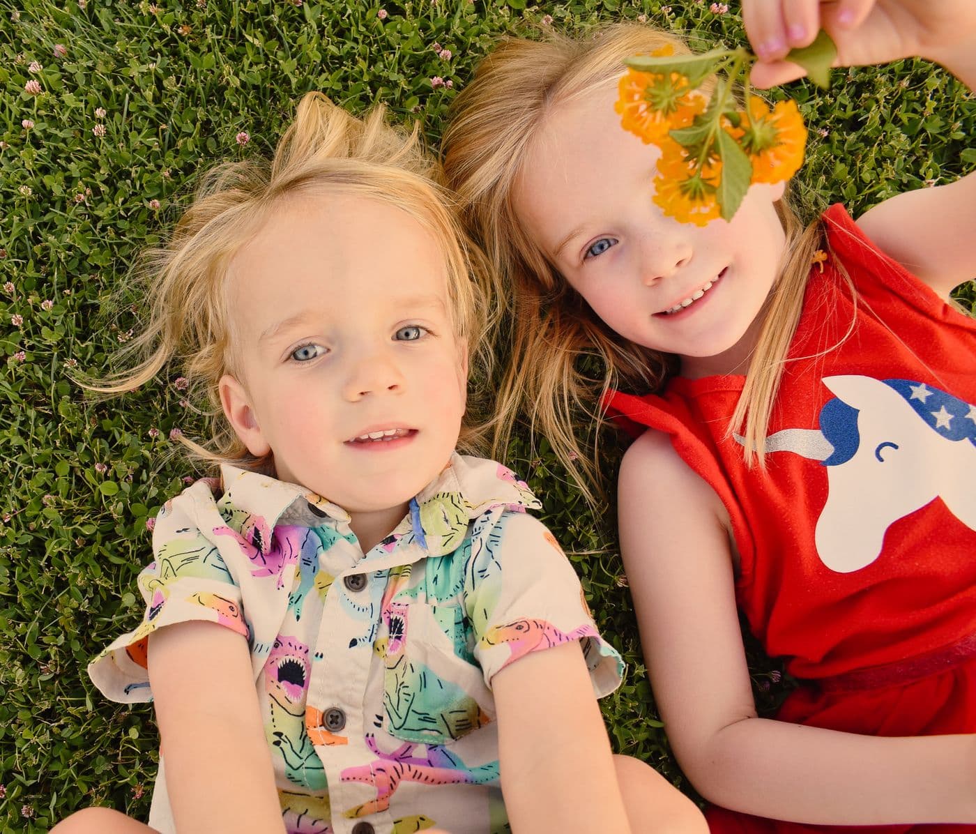 Two young children lying together in the grass during a Las Vegas family session, one holding a yellow flower to her eye