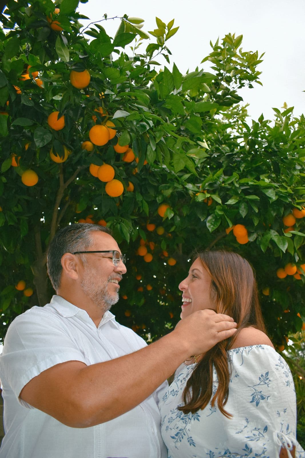 A father and his grown daughter laughing together under an orange tree during a family session in Las Vegas, NV