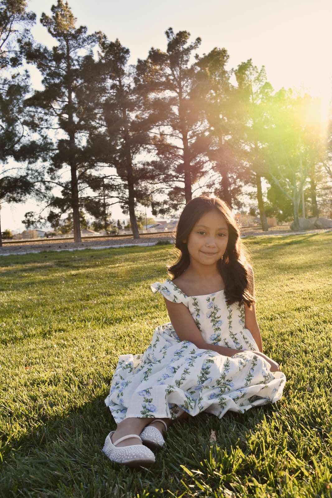 A young girl in a floral dress sitting on a sunlit lawn during a family session in Las Vegas, NV