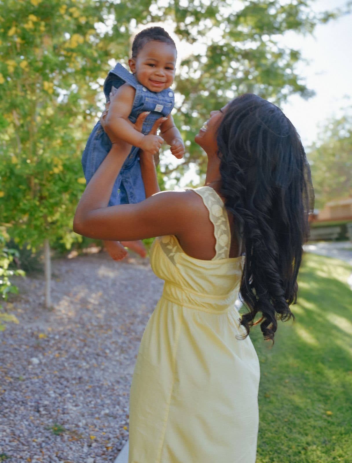A mother in a yellow dress lifting her baby up under a tree at a family photo session in Las Vegas, NV