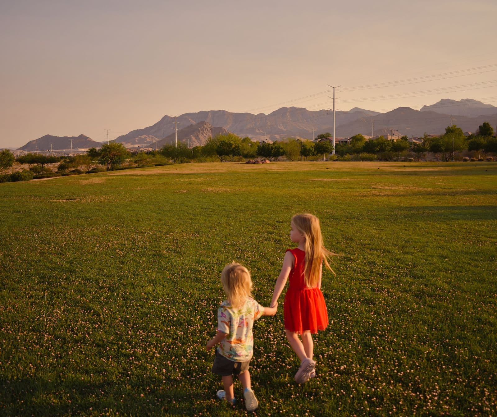 Two young siblings walking hand in hand across a lawn with mountains behind them, Las Vegas, NV