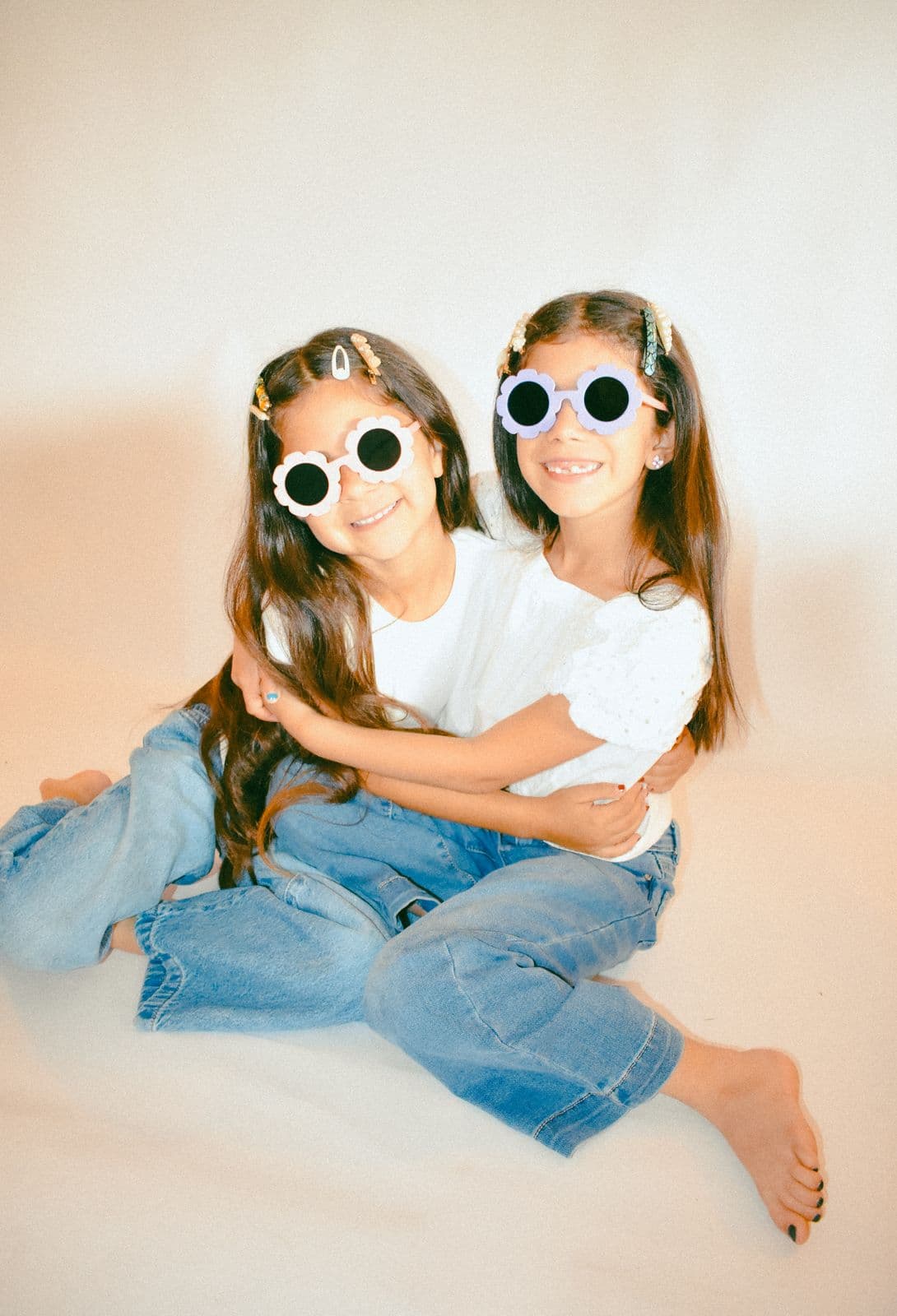 Two sisters in white tees and jeans hugging on a studio floor in round sunglasses, Las Vegas, NV