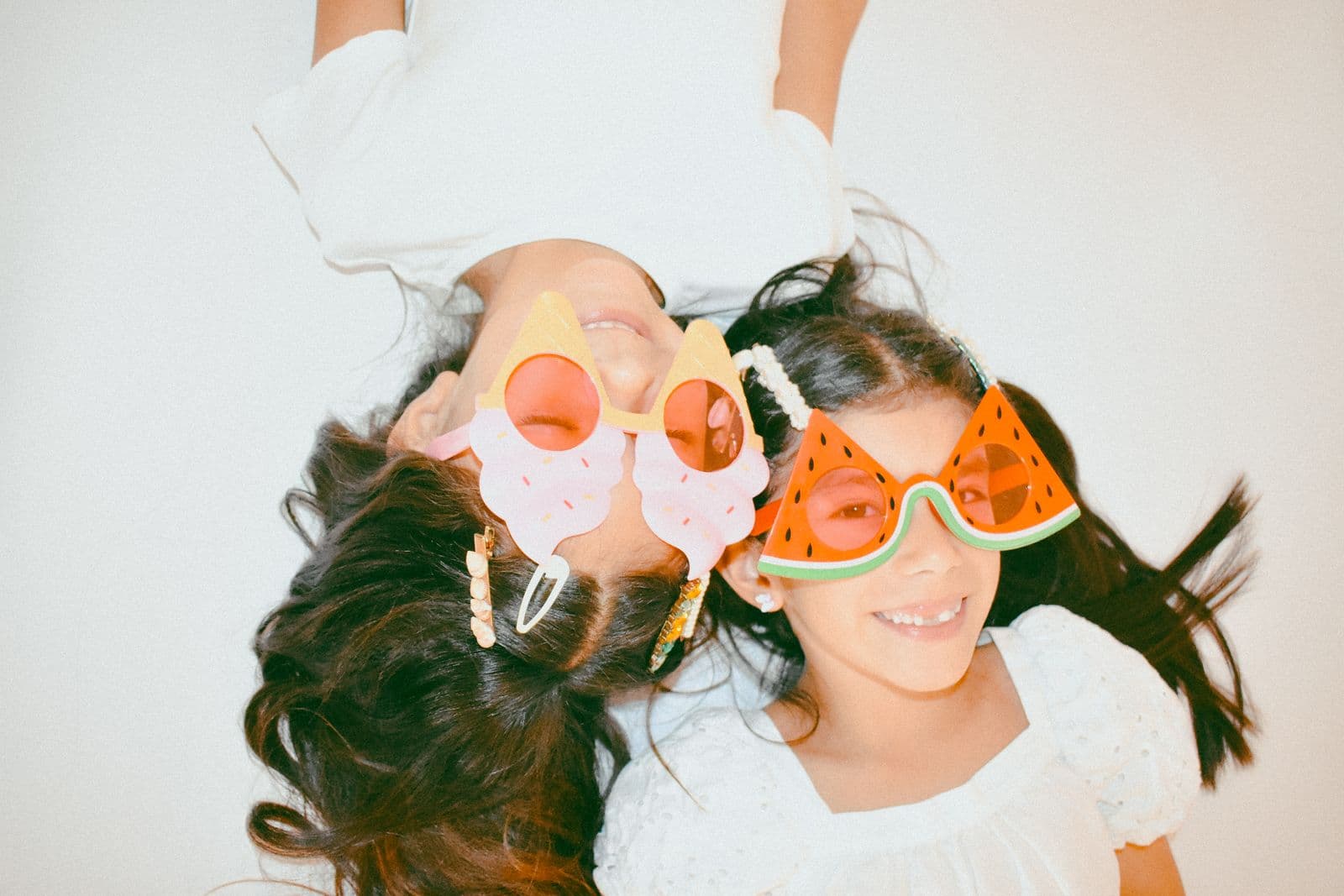 Overhead view of two sisters lying together in playful fruit-shaped sunglasses, Las Vegas, NV studio session