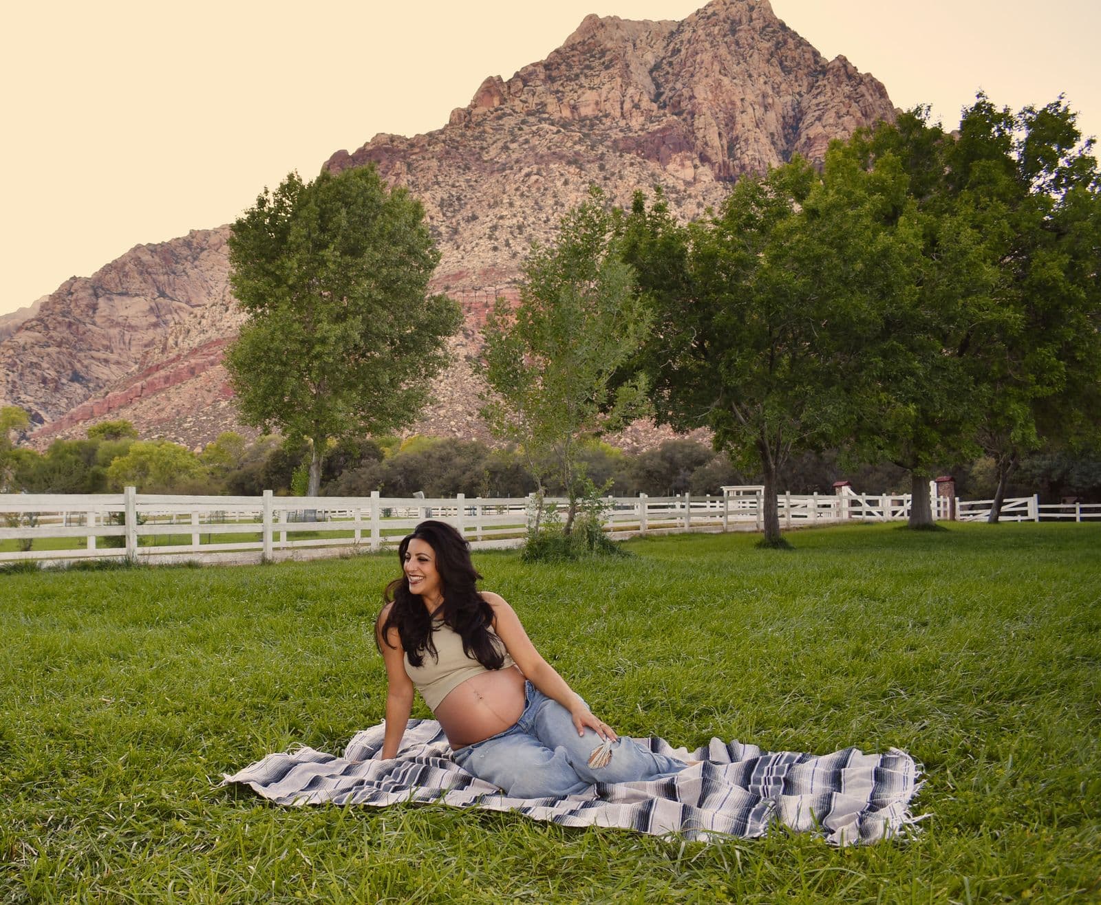 An expecting mother seated on a striped blanket on the lawn at Floyd Lamb Park in Las Vegas, cottonwood trees and desert mountains behind her