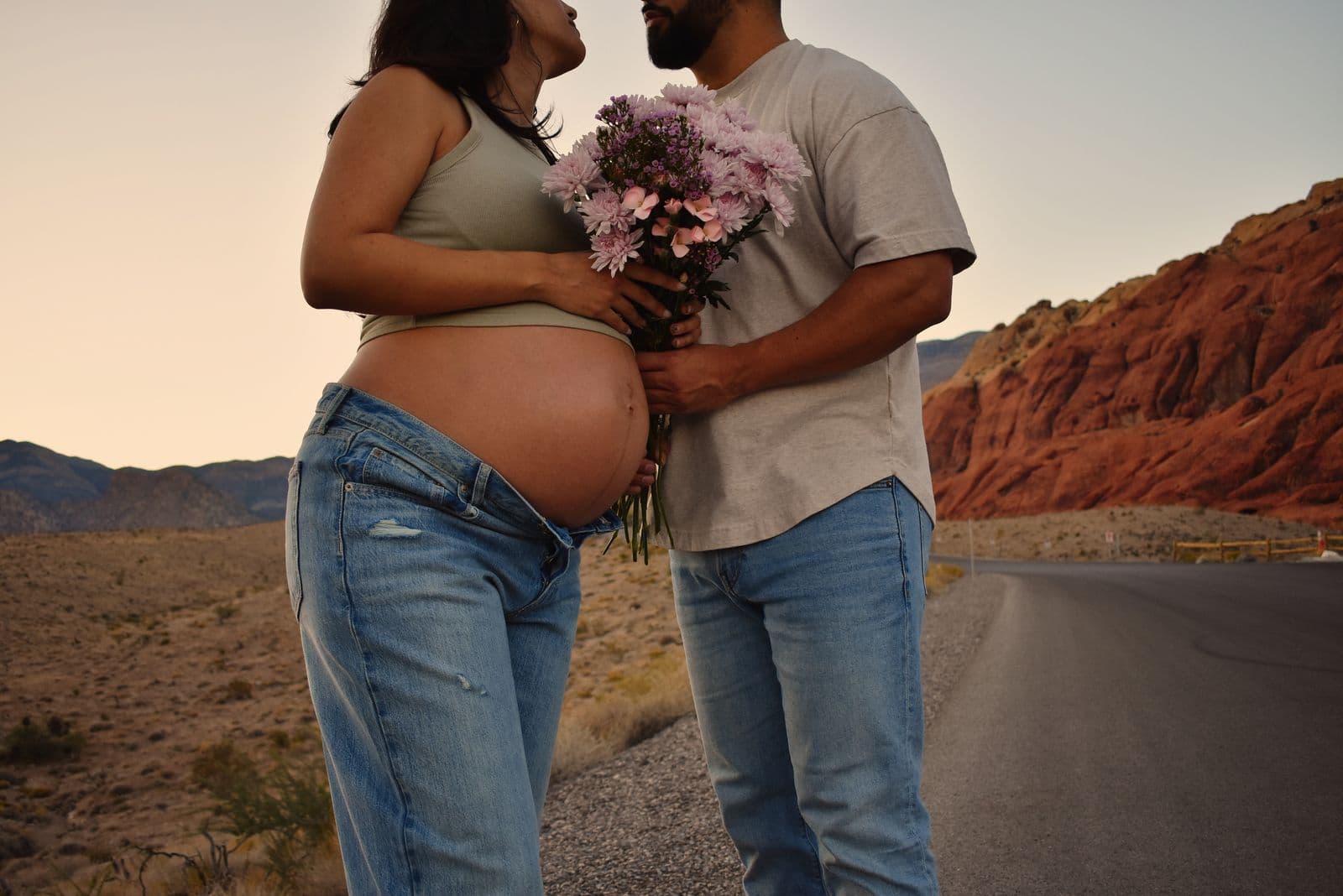 An expecting mother's bare bump beside a bouquet of pink blossoms held by her partner, framed against the red cliffs of Red Rock Canyon