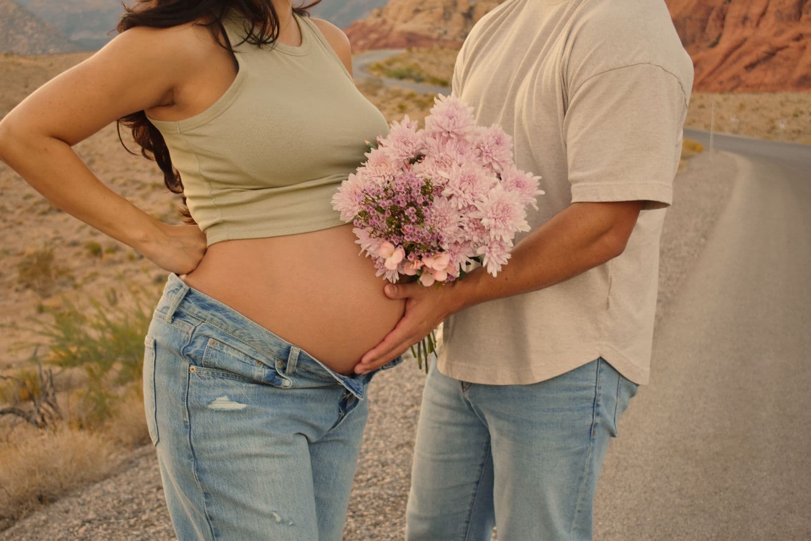 An expecting mother in a green tank top with one hand on her hip and pink flowers at her bump, photographed at Red Rock Canyon outside Las Vegas