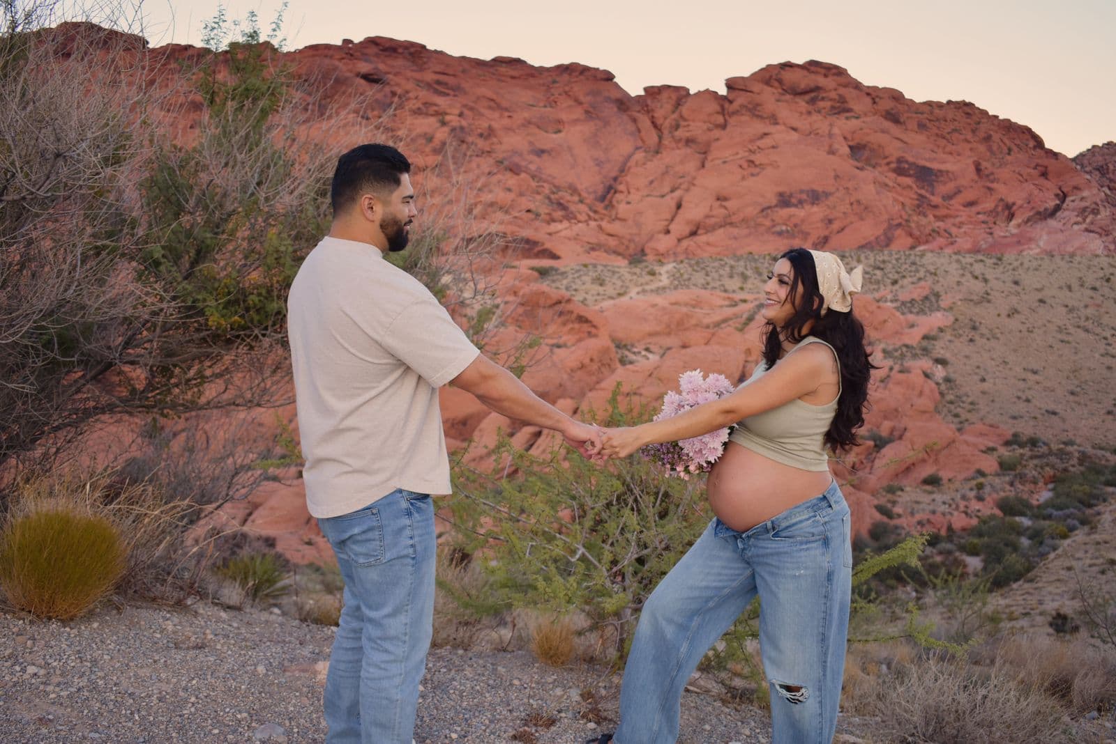 A partner reaching out to hand an expecting mother a bouquet of pink flowers in front of the red rock formations at Red Rock Canyon