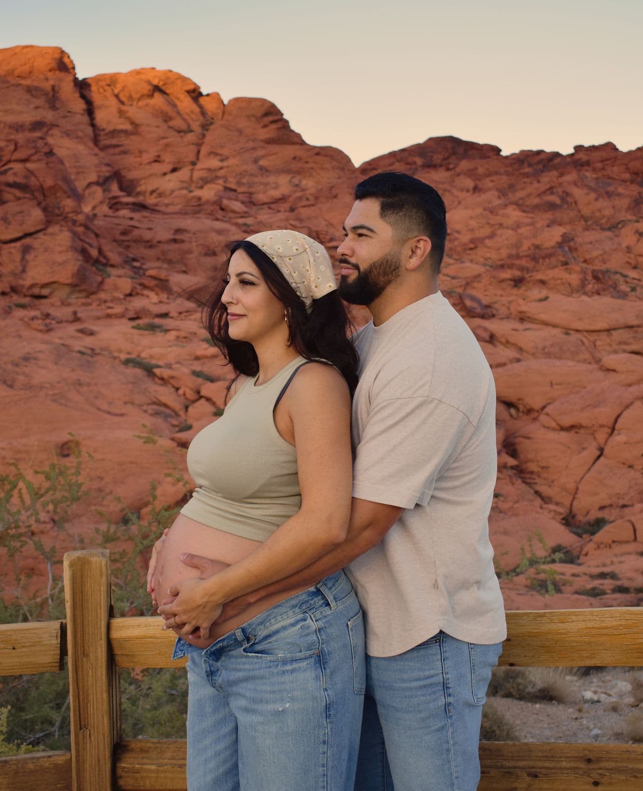 An expecting couple standing against the red sandstone of Red Rock Canyon with both of their hands resting on her bump
