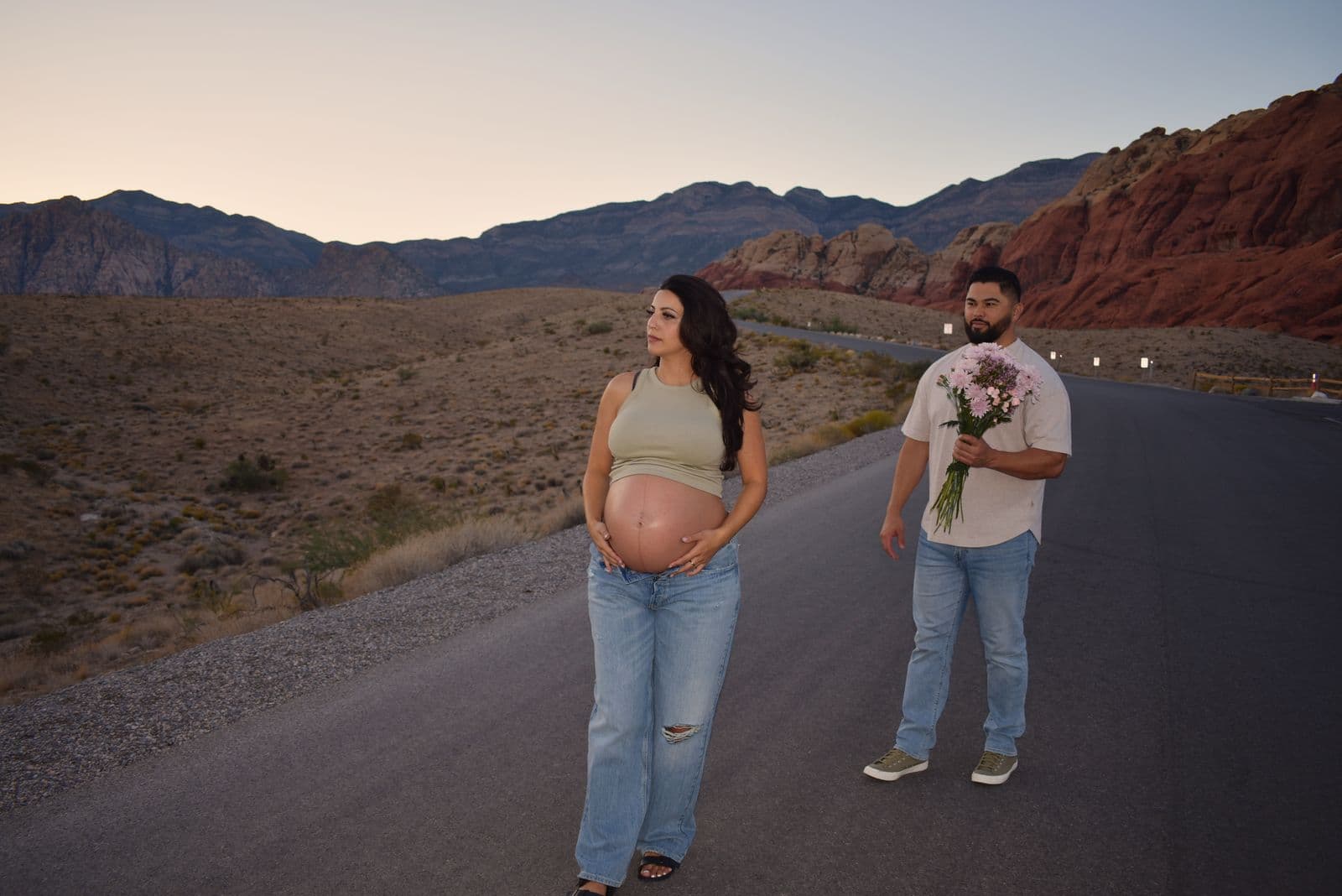 An expecting couple standing face to face on the Red Rock Canyon scenic drive at dusk, her partner holding a bouquet of pink flowers