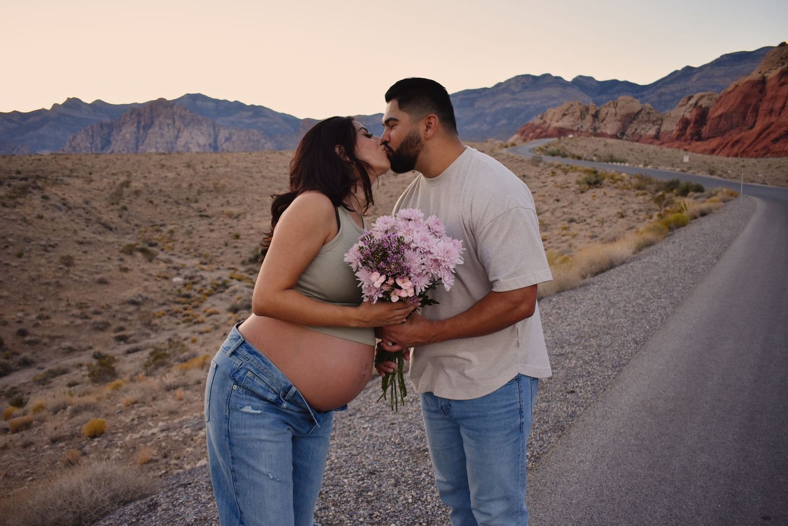 An expecting couple kissing beside the scenic drive at Red Rock Canyon near Las Vegas, a bouquet of pink flowers held between them at sunset