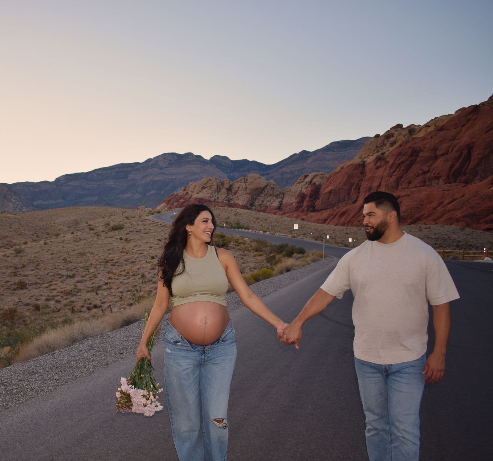 An expecting couple walking hand in hand down the Red Rock Canyon scenic drive at sunset, a bouquet of pink flowers at her side