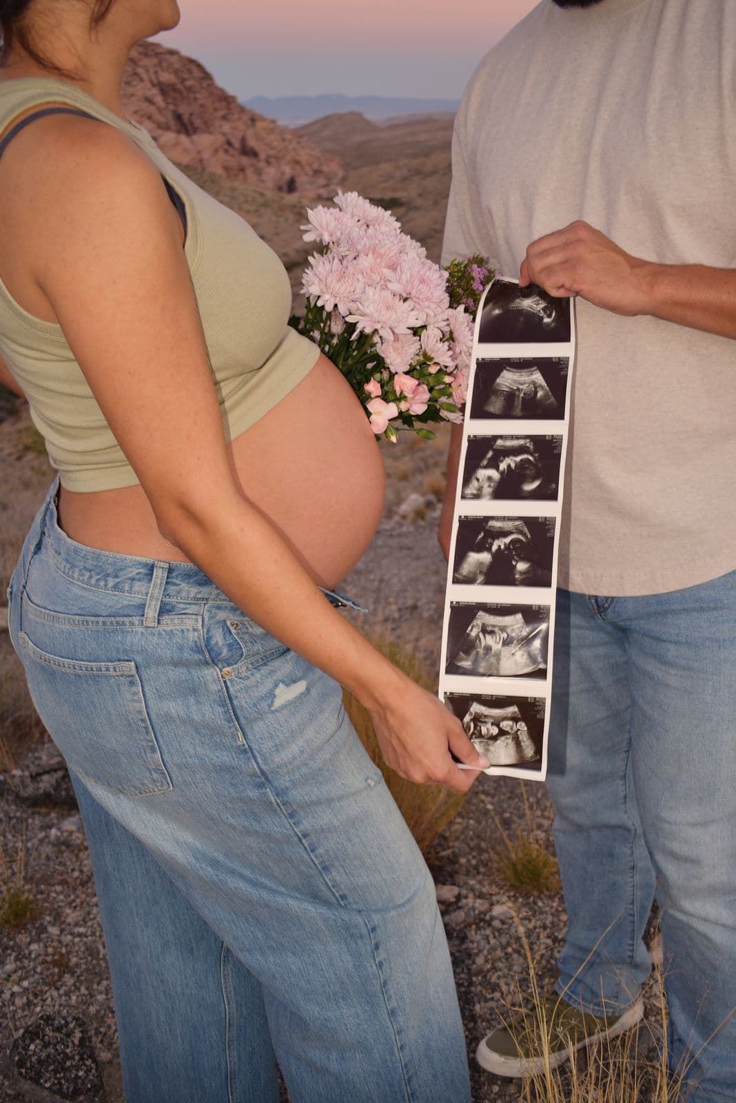 An expecting couple holding a strip of ultrasound photographs beside her bare bump at Red Rock Canyon near Las Vegas
