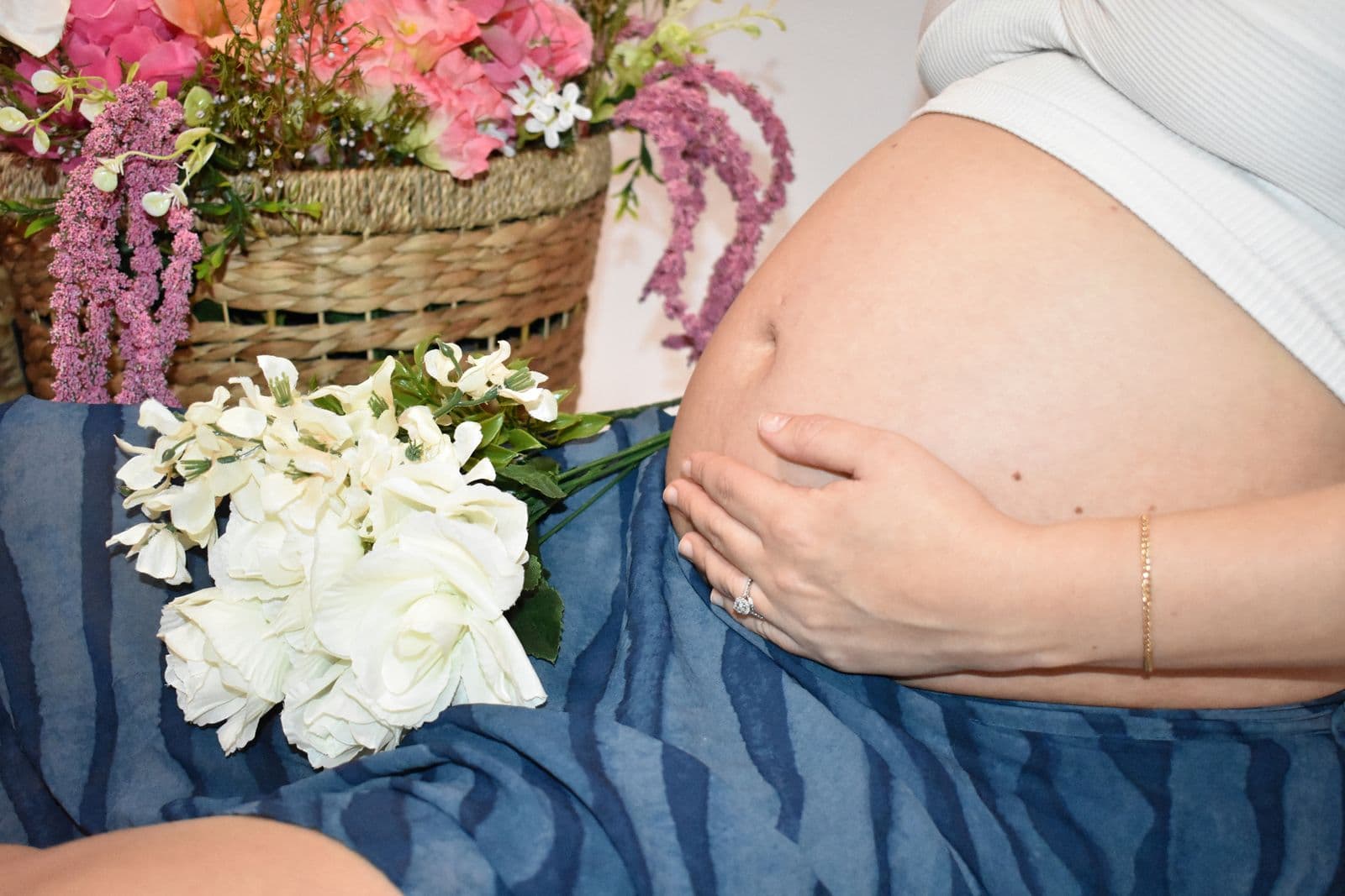 Close-up of an expecting mother's bare bump with her hand resting on it, white hydrangeas and a basket of pink flowers alongside, in a Las Vegas photography studio