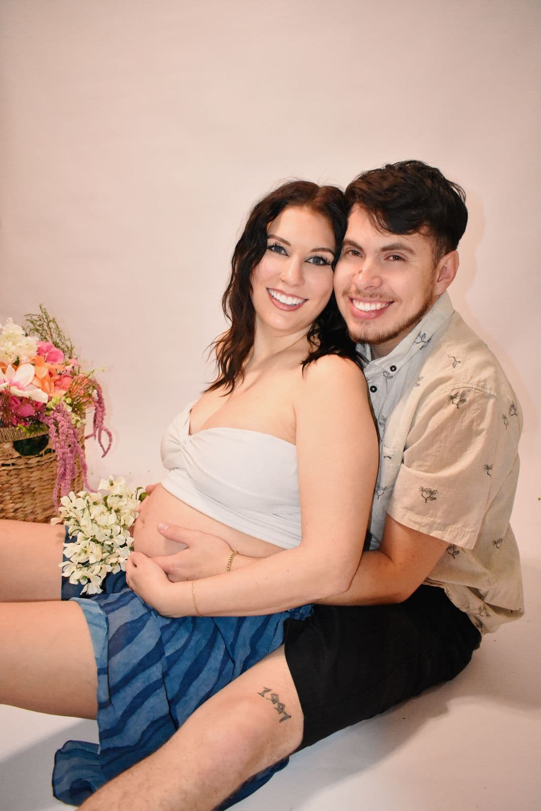An expecting couple seated together on a cream studio backdrop in Las Vegas, her partner behind her with his arms around her bare bump and a basket of pink flowers beside them