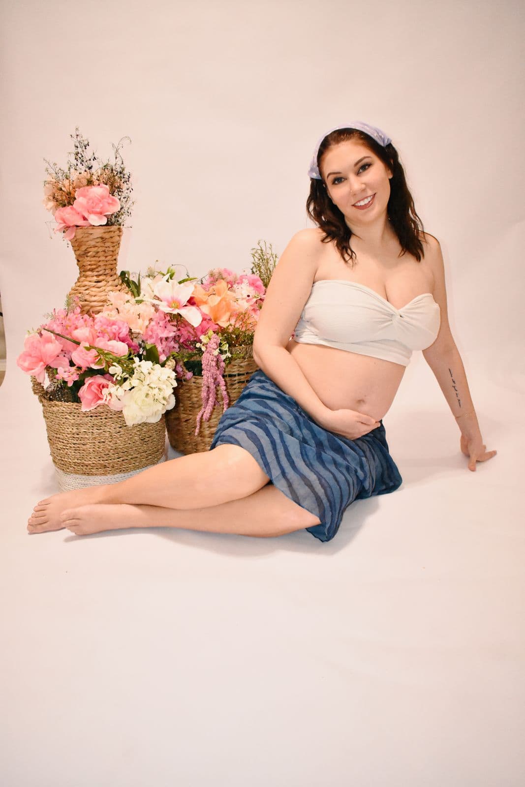 An expecting mother seated on the floor of a Las Vegas photography studio wearing a white headband, two woven baskets of pink and peach flowers beside her