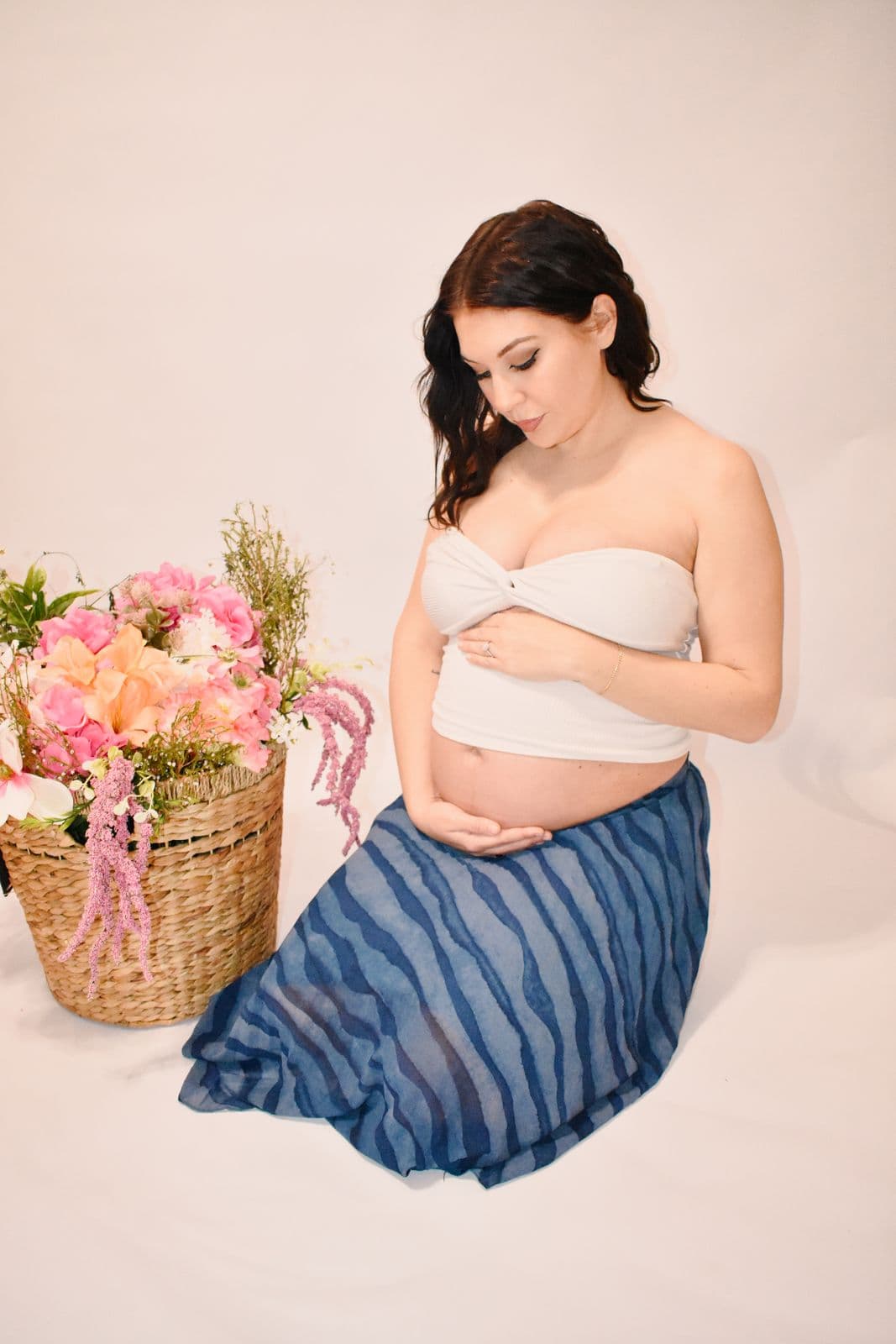 An expecting mother seated on a cream studio backdrop in Las Vegas, looking down at her bump with both hands resting on it