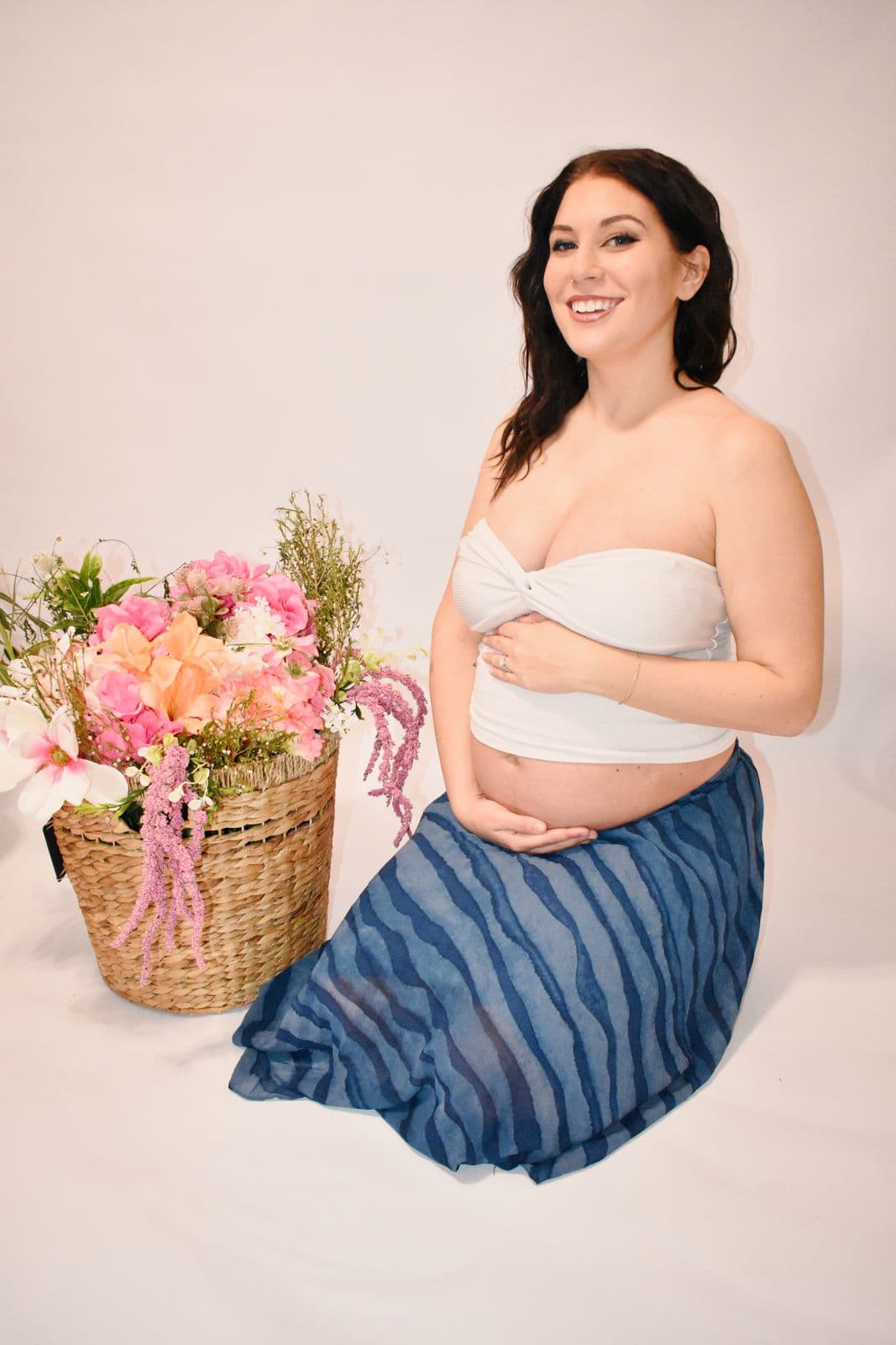 An expecting mother smiling at the camera during a Las Vegas studio maternity session, one hand on her bump beside a woven basket of pink and peach flowers