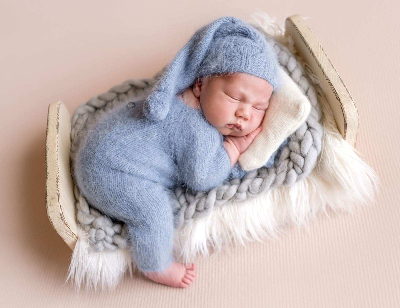 A newborn in a blue knit romper and sleepy cap curled in a small cream bed on a soft pink backdrop, in studio in Las Vegas, NV