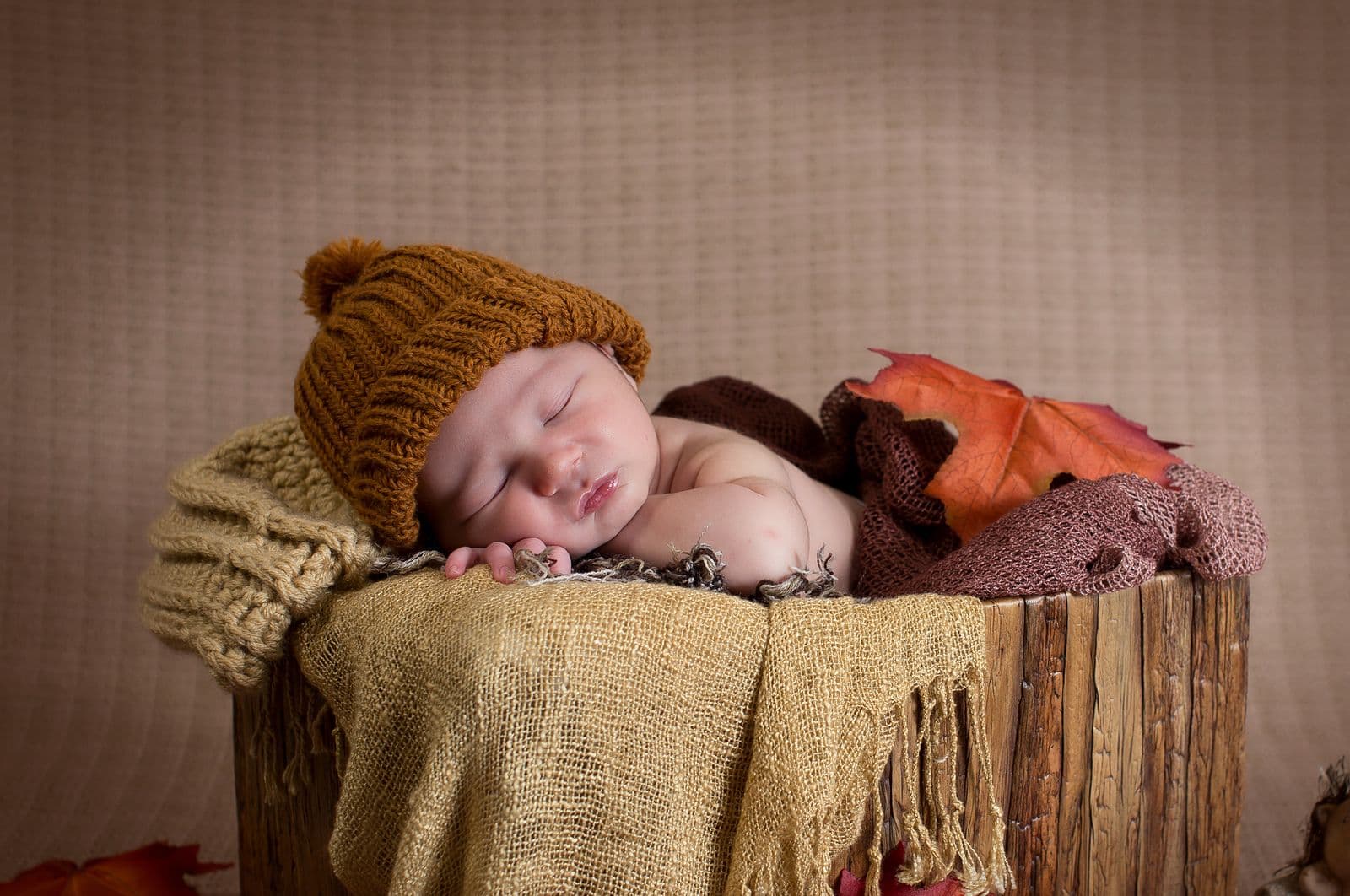 A newborn in a mustard knit hat asleep in a burlap-lined wooden bucket with fall leaves, in studio in Las Vegas, NV