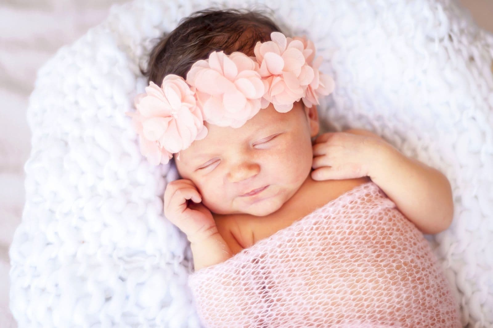 A newborn girl in a peach floral headband and pink wrap asleep on a chunky white knit blanket, in studio in Las Vegas, NV