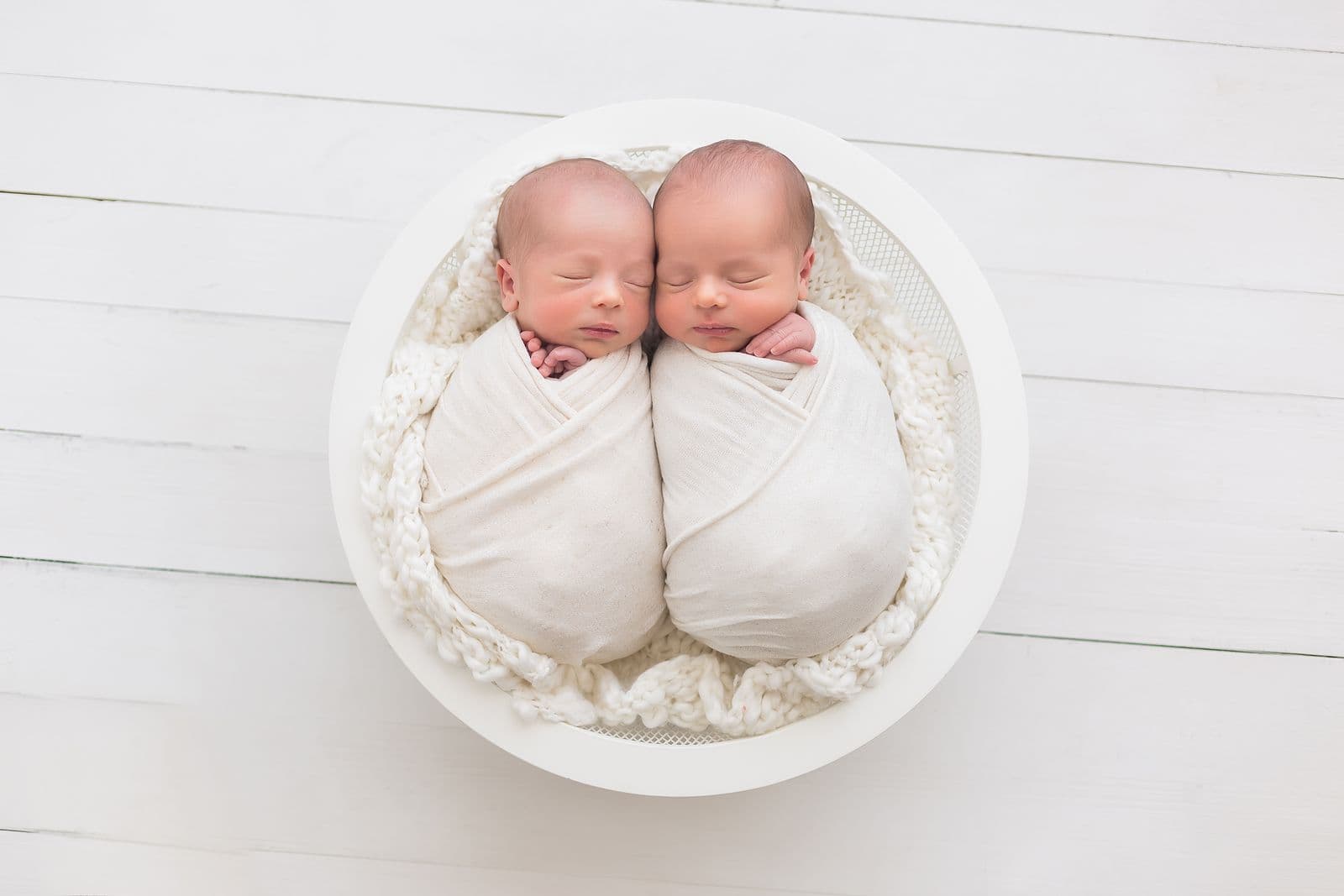 Newborn twins swaddled in white and nestled together in a round bowl on a white wood floor, in studio in Las Vegas, NV