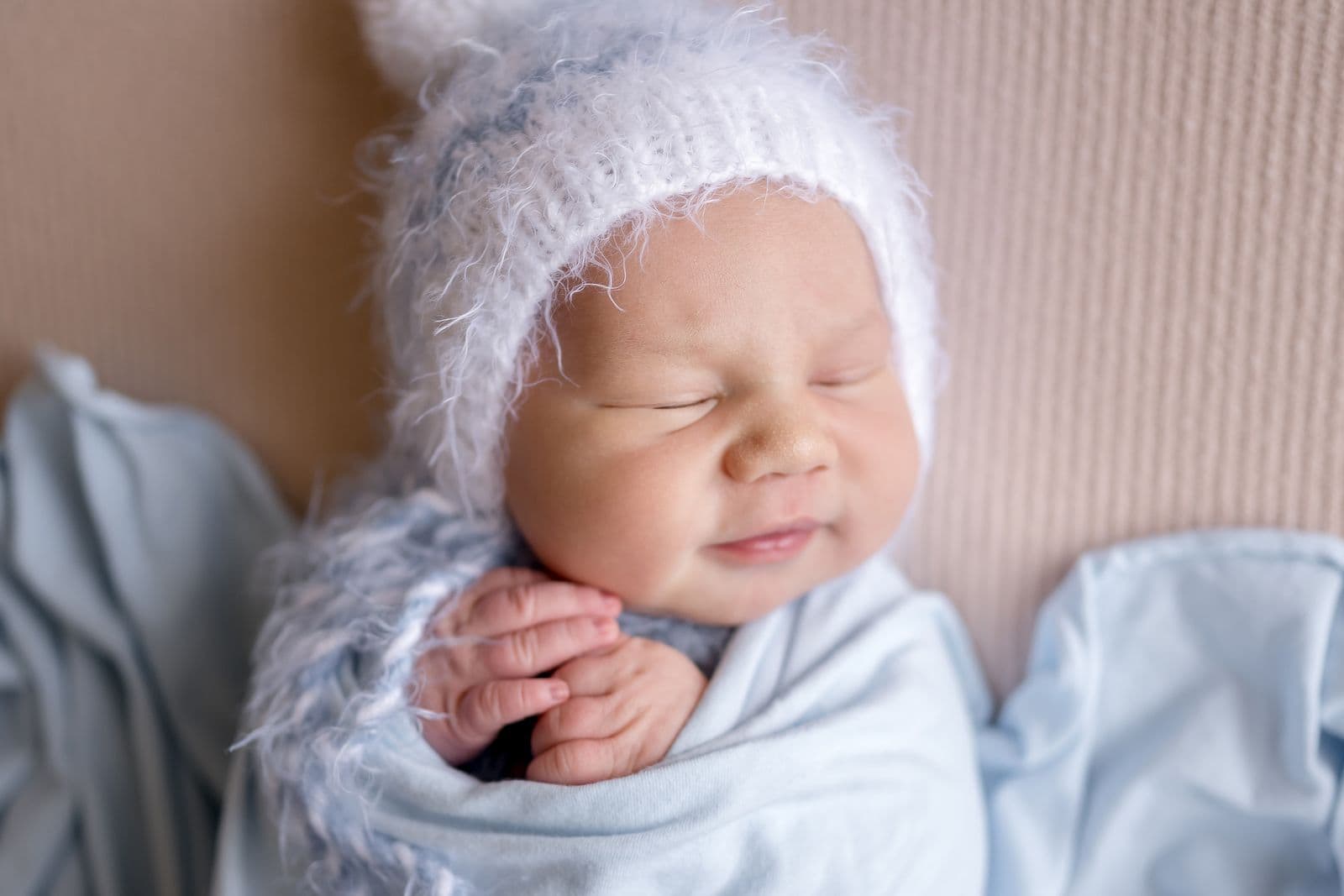A newborn in a fluffy white bonnet wrapped in pale blue with both hands tucked under one cheek, in studio in Las Vegas, NV