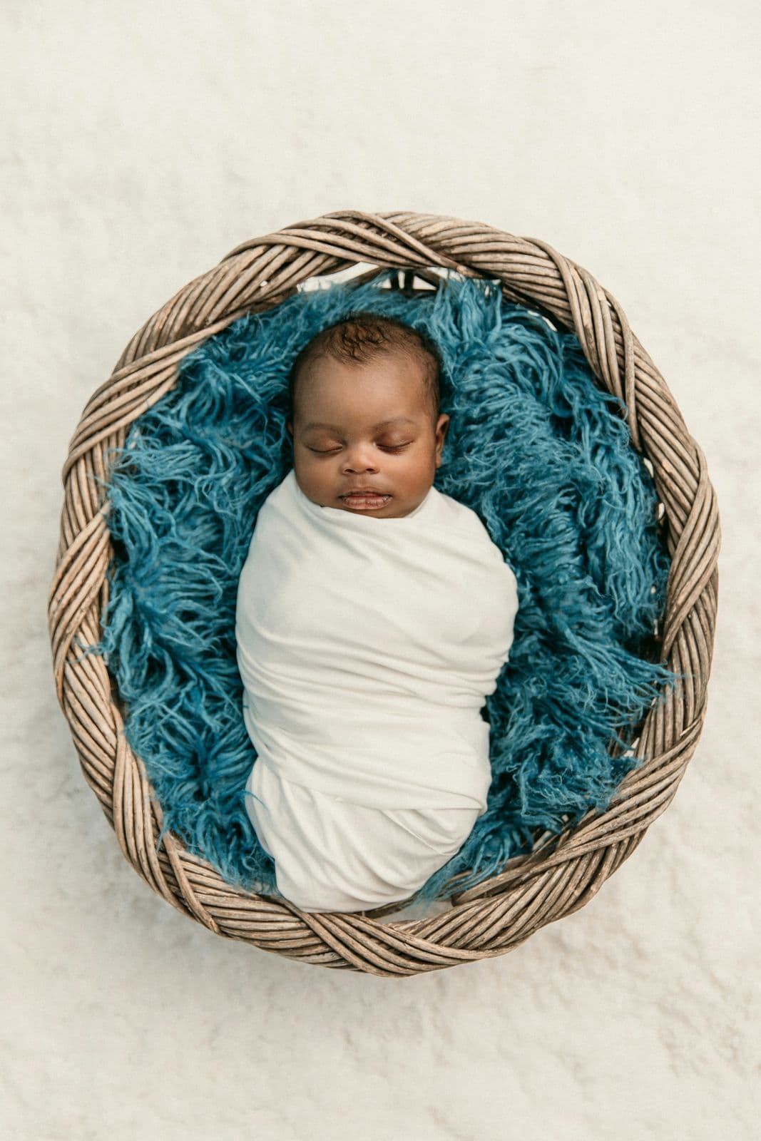 A newborn swaddled in white and resting in a round woven basket lined with teal fur, in studio in Las Vegas, NV