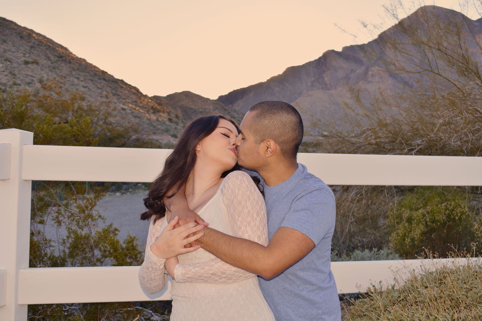 A couple kissing beside a white ranch fence with desert mountains behind them at sunset near Las Vegas