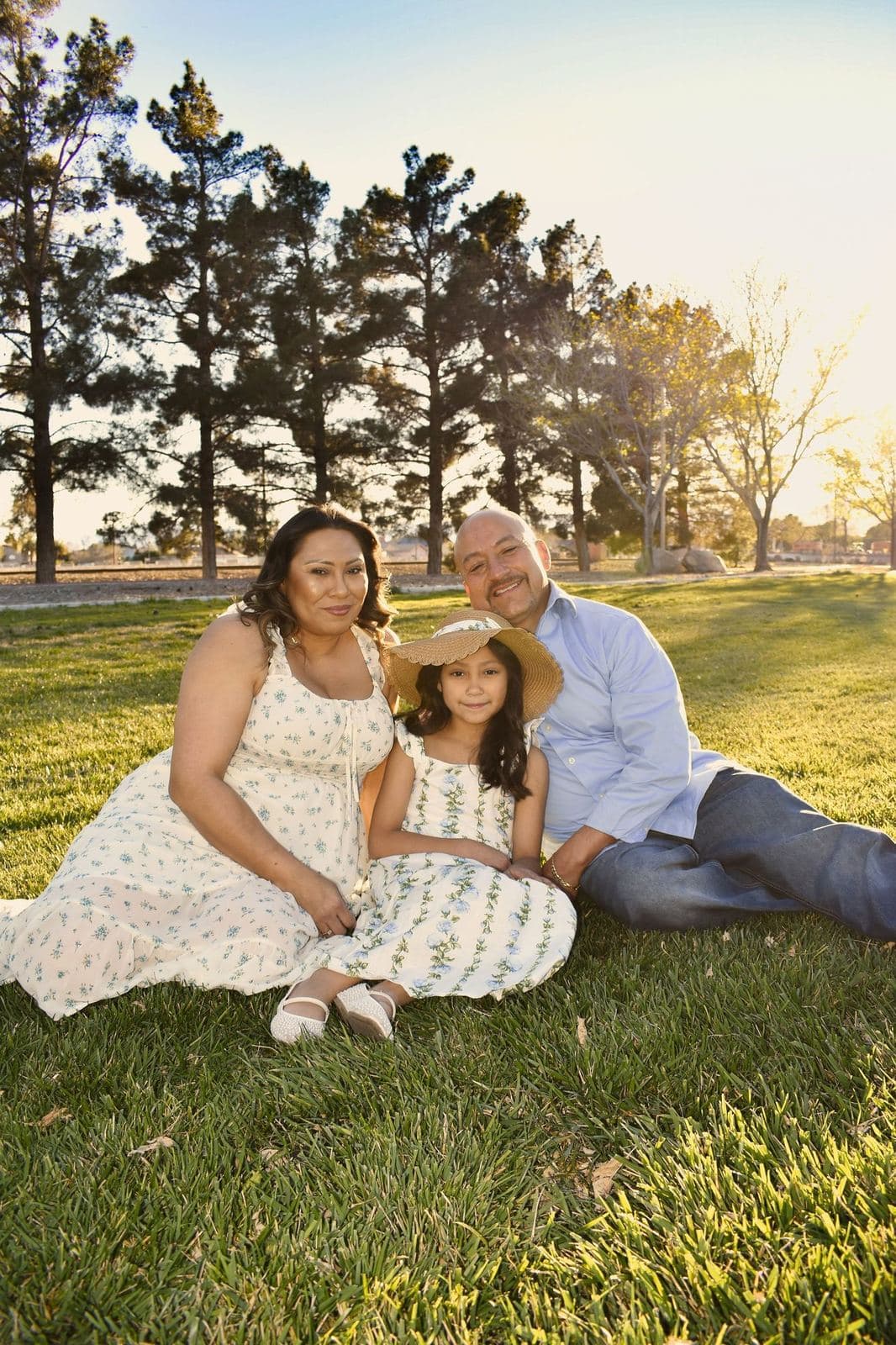 A family of three sitting on green grass in a sunny Las Vegas park, parents smiling with a young girl in a straw hat between them