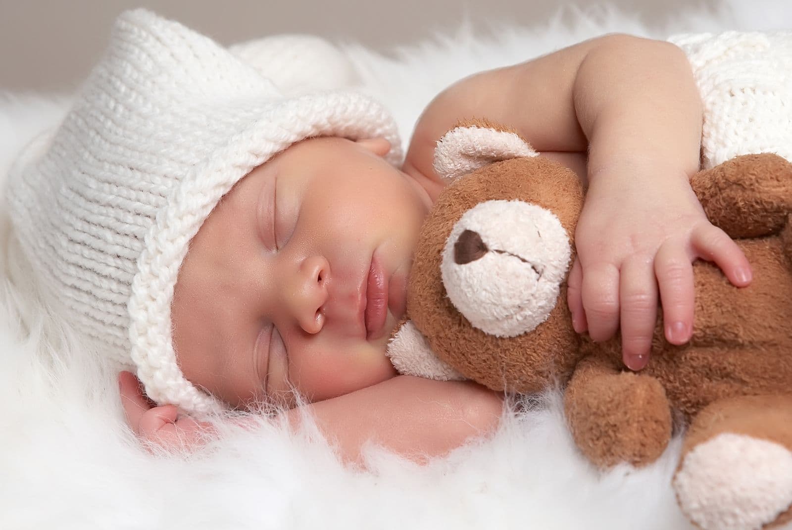 A newborn in a cream knit cap asleep with one arm around a teddy bear during an in-studio newborn session in Las Vegas, NV